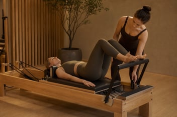 Mujer haciendo footwork en un reformer de madera con una instructora ajustando la resistencia